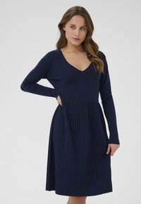 Femme aux longs cheveux portant une robe bleu marine avec un décolleté en V et une jupe plissée, posant avec une main sur la hanche devant un fond uni.