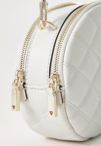Borsa a tracolla bianca in similpelle con motivo trapuntato, doppie cerniere dorate e dettagli quadrati. Presenta una forma arrotondata e una texture liscia.