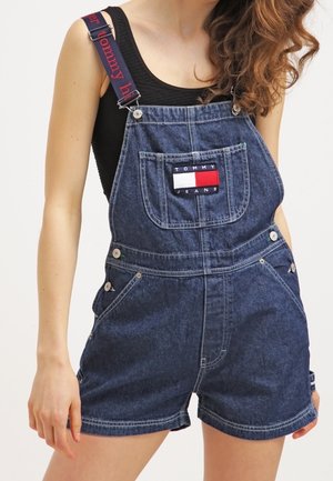 Kvinde iført mørkeblå denim overall shorts med Tommy Jeans-logo og sort ærmeløs top, med håret løst på den ene side.