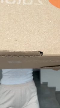 Scatola di cartone marrone con una superficie liscia, presenta un angolo ritagliato. La parte superiore mostra un logo bianco, con un accento arancione circolare.