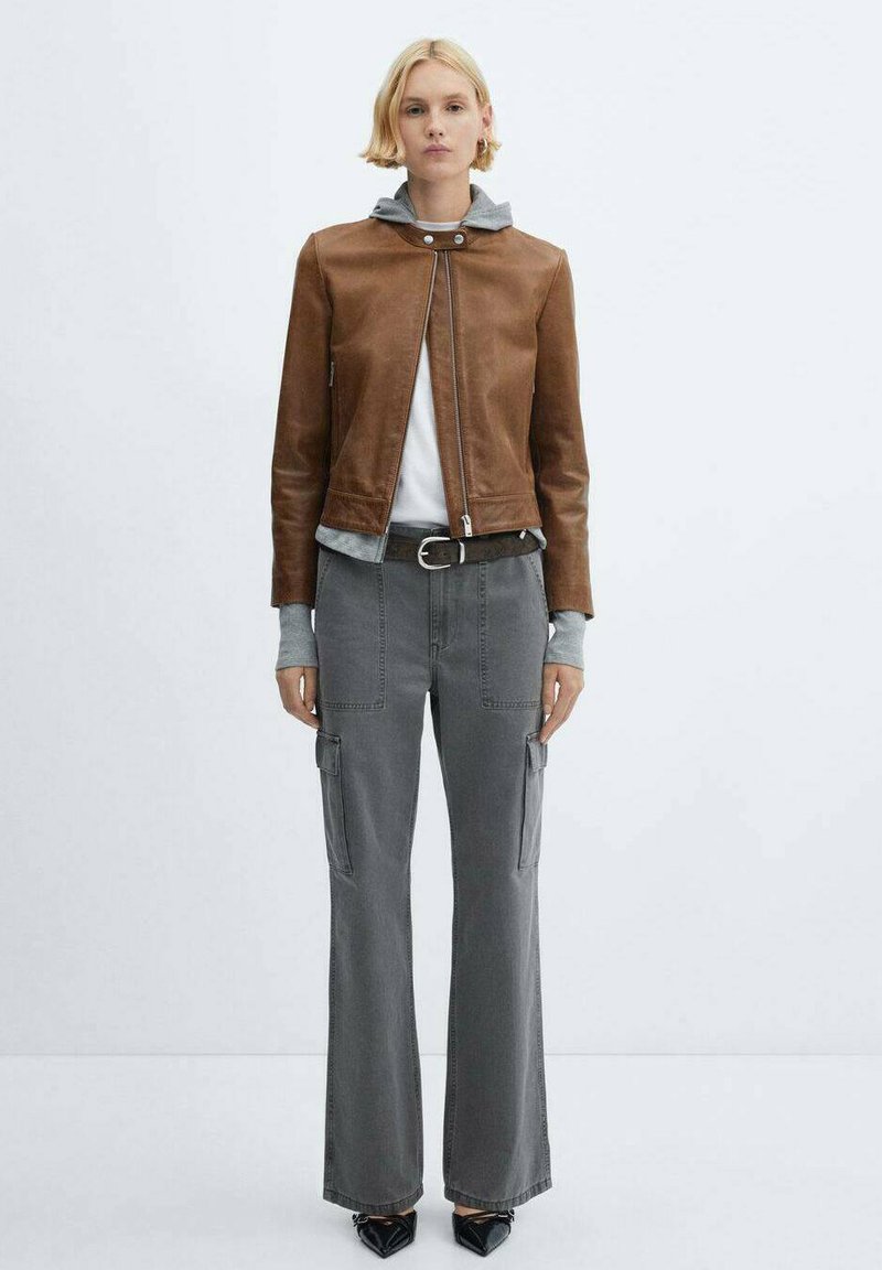 Brązowa skórzana kurtka z zamkiem, szara bluza z kapturem pod spodem, szare spodnie cargo i czarne buty w szpic. Minimalistyczny design z kieszeniami użytkowymi.