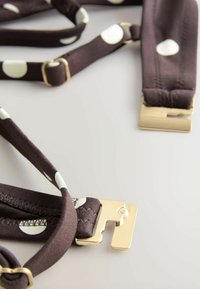 Suspenders di stoffa marrone decorate con pois bianchi. Presentano una minuteria in metallo dorato con una clip rettangolare e dettagli di cucitura strutturata.
