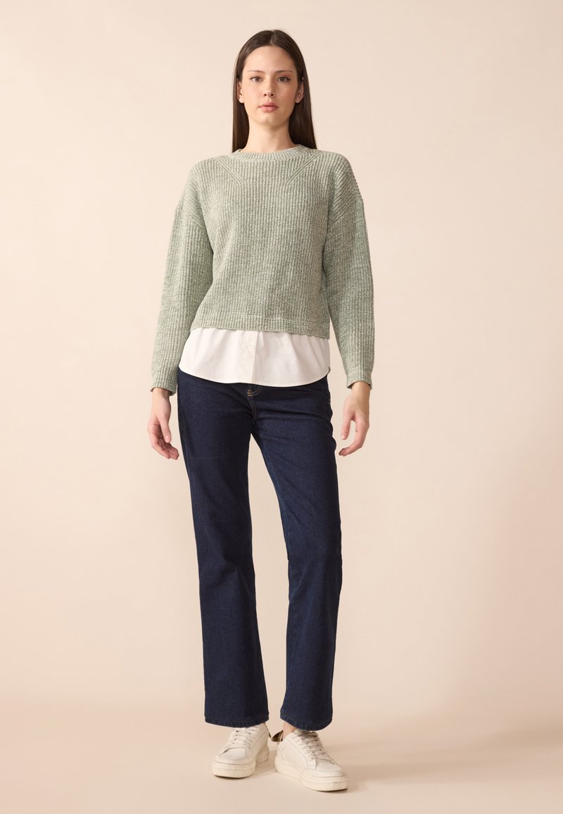 Pull en maille vert clair avec un motif texturé, porté par-dessus une chemise blanche à col, associé à un jean bleu foncé à coupe droite et des baskets blanches.
