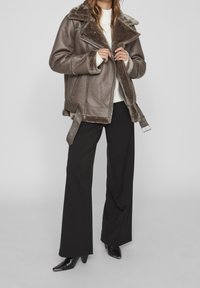 Giacca in pelle marrone con foderatura in pelliccia, colletto oversize, chiusura con zip e dettagli con fibbie sui lati, abbinata a pantaloni neri a gamba larga e scarpe.