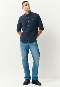 Hombre de pie con las manos detrás de la espalda, vistiendo una camisa de botones azul marino, jeans de un azul claro y zapatos negros, contra un fondo liso.