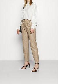 Beige Lederhosen mit Falten und Nahtdetails, kombiniert mit einer weißen Bluse mit hohem Kragen und weit schwingenden Ärmeln. Schwarze Sandalen mit Absatz.