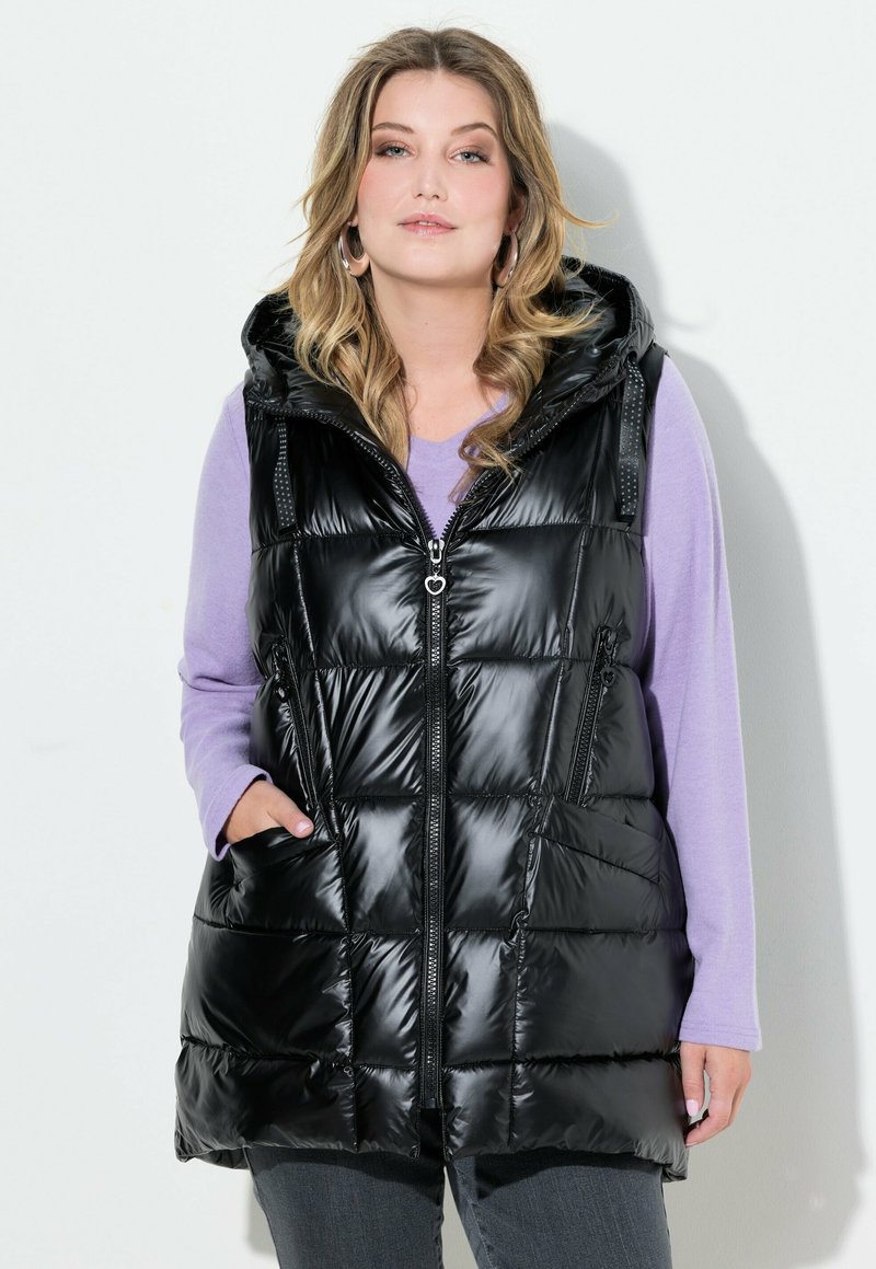 Schwarze gesteppte Weste mit Kapuze, mit Reißverschluss vorne und seitlichen Taschen. Getragen über einem helllila Langarmshirt.