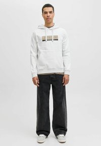 Lichtgrijze hoodie met contrasterend logo op de borst, gecombineerd met losse zwarte jeans en lichtgrijze schoenen. Minimalistisch ontwerp, casual stijl.