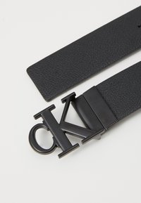 Schwarzer Ledergürtel mit einer strukturierten Oberfläche und einer großen schwarzen Metallschnalle in Form von "CK". Die Schnalle hat ein minimalistisches Design.