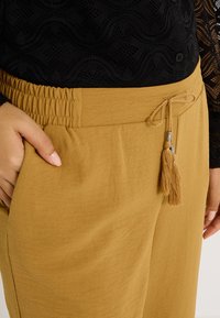 Pantalons marron clair avec une taille élastique et un cordon de serrage ajustable noué avec des grelots. Le tissu semble doux avec une texture lisse.