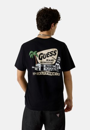 Uomo con maglietta nera con grafica in stile retrò "Guess Jeans Hotel & Restaurant from California" con palma e diner.