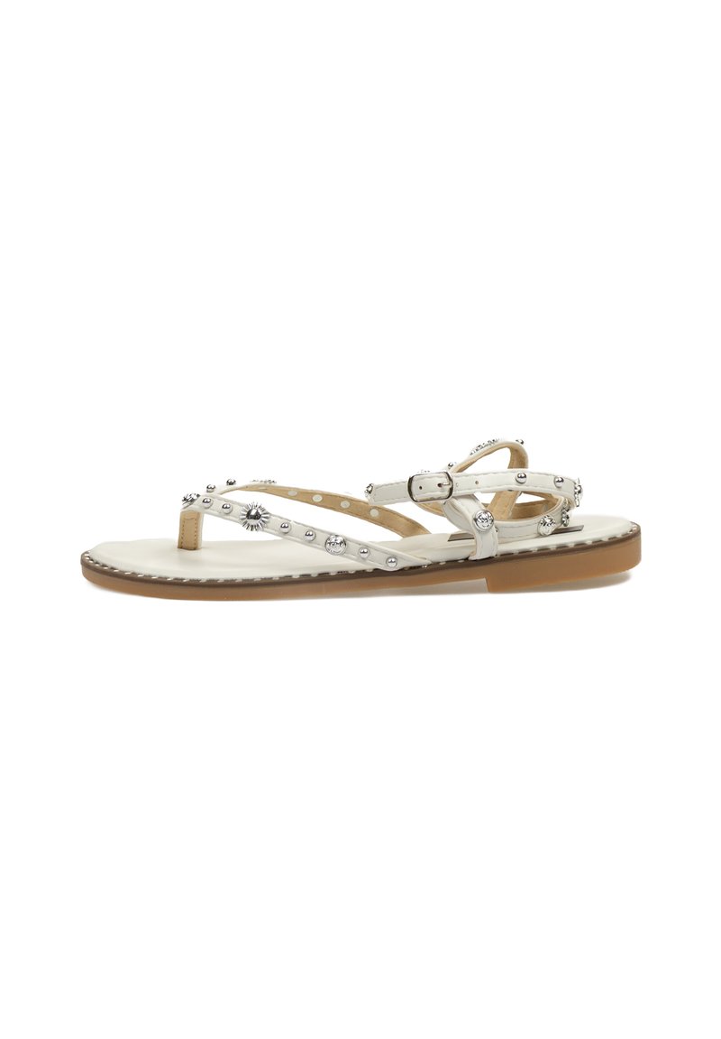 Sandalias planas blancas con tachuelas plateadas, que presentan un diseño minimalista, correa en el tobillo ajustable y una suela texturizada beige.
