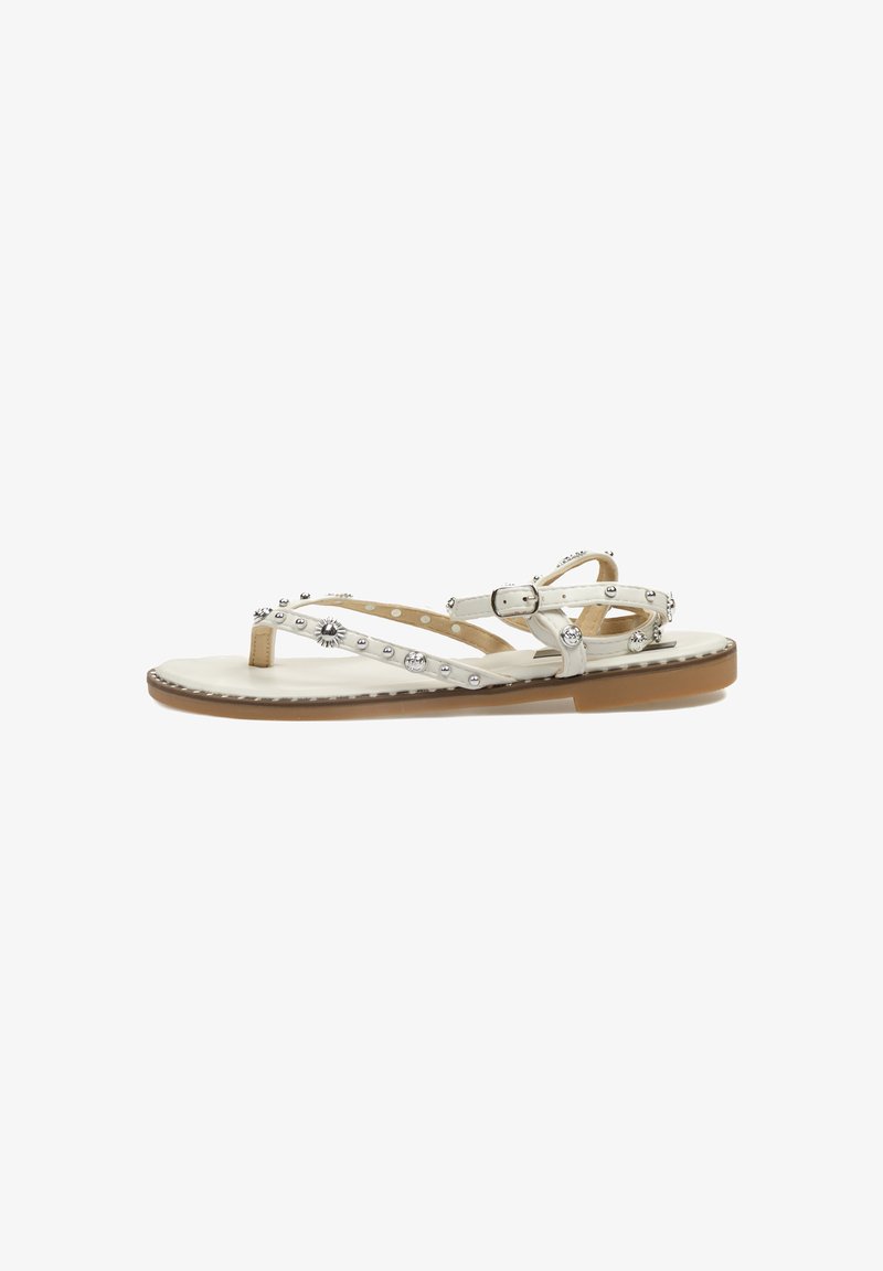 Sandalias planas blancas con tachuelas plateadas, que presentan un diseño minimalista, correa en el tobillo ajustable y una suela texturizada beige.