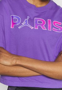 Fialové zkrácené tričko s odvážným, vícebarevným nápisem "PARIS" a siluetou Jumpman. Krátké rukávy, hladká bavlněná tkanina, volný střih.