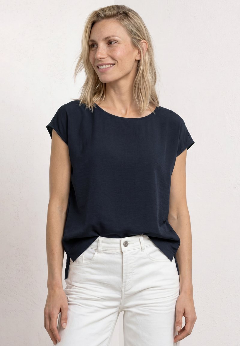 Femme blonde souriante regardant vers la gauche, portant un haut ample bleu marine foncé à manches courtes et un pantalon blanc taille haute sur un fond clair.