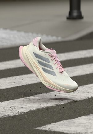 Light-colored running shoe with pink accents, textured upper, three silver stripes, and a cushioned sole, shown in mid-air over a crosswalk.