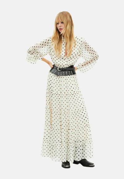 Langes weißes Kleid mit schwarzen Herzmustern, transparentem Stoff, aufgeblähten Ärmeln, geknöpftem Kragen und breitem schwarzen Gürtel mit silberner Beschläge. Schwarze Stiefel.