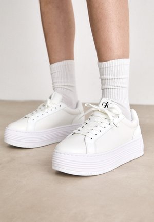 Sneakers platform bianche realizzate in pelle liscia, con una spessa suola bianca dotata di righe orizzontali e design con lacci. Dettaglio del logo nero.