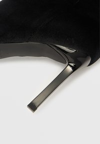 Fekete velúr magassarkú cipő letisztult kialakítással, hegyes orral és fényes, fekete tűsarokkal. Sima textúra és elegáns forma.