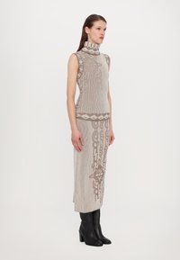 Robe en tricot beige avec des accents à motifs ; col roulé haut, design sans manches et jupe mi-longue, assortie à des bottes à talons noires.