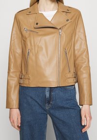 Femme portant une veste de moto en simili cuir beige avec des zips et des boucles argentés, associée à un jean en denim taille haute bleu.