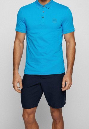 Mężczyzna ubrany w jasnoniebieską koszulkę polo z krótkim rękawem z trzema guzikami i granatowe szorty, stojący na jednolitym tle.