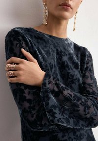 Vrouw met doorschijnende zwarte fluwelen top met bloemenpatroon, gouden oorbellen en meerdere gestapelde gouden ringen aan haar vingers, armen over elkaar geslagen op haar borst.