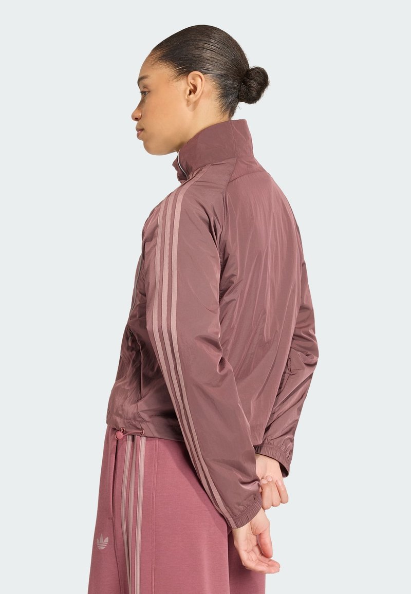 Femme aux cheveux attachés en chignon portant une veste de sport mauve et un pantalon assorti avec trois bandes claires sur les manches et les jambes, debout de profil.