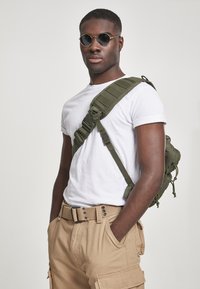 Olīvu zaļš taktiskais mugursomas, nēsāts uz šķērsu, apvienots ar pieguļošu baltu T-kreklu un bēša krāsas kraukšķīgiem šortiem, kas aprīkoti ar kabatām un auduma jostu.