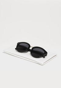 Ochelari de soare negri în stil cat-eye cu lentile închise. Ramă lucioasă, detaliu auriu la balama, așezați pe o etui albă, texturată.