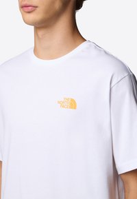 Valkoinen puuvillapaita, jossa on pieni keltainen "The North Face" -logo vasemmassa rinnassa. Paidassa on klassinen pyöreä kaula-aukko ja lyhyet hihat.