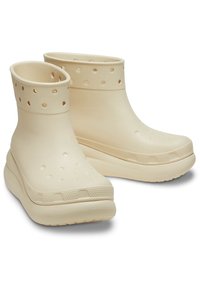 Bottes en caoutchouc beige jusqu'à la cheville avec une finition lisse, des perforations circulaires et une base texturée. Elles disposent d'un bout arrondi rembourré et d'une semelle épaisse.