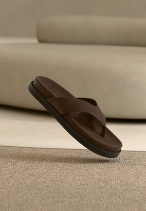 Braune Lederflipflop-Sandale mit schwarzer Sohle, die über einem beigen Teppich in einer neutraltonigen Inneneinrichtung schwebt.
