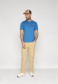 Μπλε κοντομάνικο μπλουζάκι με γραφικό βουνό και κείμενο "patagonia". Μπεζ παντελόνια με φερμουάρ που αφαιρούνται και μπεζ καπέλο με πλέγμα στην πίσω πλευρά. Άσπρα αθλητικά παπούτσια.