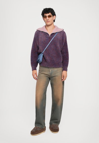 Lila und rosa gestrickter Pullover mit einem Viertelreißverschluss, kombiniert mit hellen, ausgewaschenen Baggy-Jeans und braunen Slippern. Blaue Umhängetasche und Sonnenbrille.