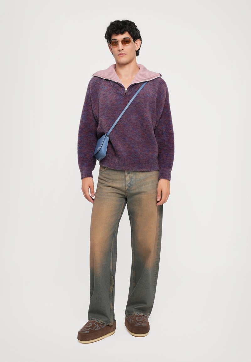 Purple and pink knitted sweater with a quarter zip, paired with light faded baggy jeans and brown slip-on shoes. Blue crossbody bag and sunglasses.