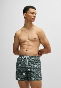 Green swim shorts featuring a repeating bold black and white "HUGO" pattern, with an elastic waistband and white drawstring detail.