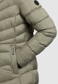 Puffer jacket in light olive green with quilted design, featuring a zippered pocket and a round logo patch on the sleeve.