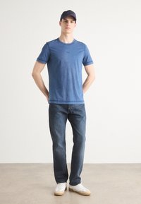 Μπλε βαμβακερό t-shirt με κοντά μανίκια, στρογγυλή λαιμόκοψη και μια διακριτική λογότυπο; συνδυασμένο με σκούρες τζιν παντελόνες και λευκά αθλητικά παπούτσια με μπεζ σόλες.