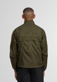 Muž s krátkými vlasy, oblečený v olivově zelené bundě s jemným textem na zádech, stojící proti jednobarevnému světle šedému pozadí.