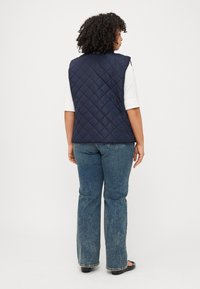 Persona con capelli ricci di spalle, che indossa un gilet trapuntato blu navy, una maglia bianca e jeans svasati, è in piedi davanti a uno sfondo bianco semplice.