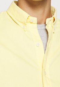 Žlutá košile na knoflíky s texturovanou, měkkou látkou. Má límec zapínaný na knoflíky a viditelnou šedou tričko pod ní.