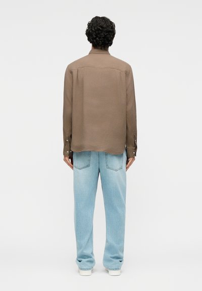 Άτομο με σγουρά σκούρα μαλλιά που φορά καφέ μακρυμάνικο πουκάμισο και ανοιχτόχρωμο μπλε τζιν, στέκεται με την πλάτη προς τα εμάς, μπροστά σε λευκό φόντο.