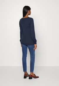 Top azul marinho de mangas longas com decote redondo, combinado com jeans ajustados de denim azul e mocassins de salto castanhos, visto de trás.