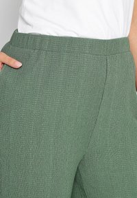 Texturované zelené kalhoty s elastickým pasem, s jemným žebrovaným vzorem a postranními kapsami, zobrazené proti bílému topu.