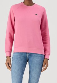 Różowa bluza z okrągłym dekoltem, ściągaczami na mankietach i u dołu. Na piersi małe zielone logo i teksturowana tkanina. W zestawie z niebieskimi dżinsami.