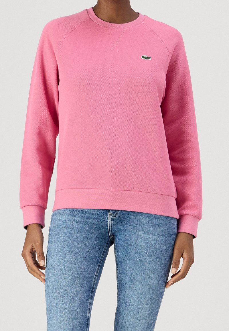 Różowa bluza z okrągłym dekoltem, ściągaczami na mankietach i u dołu. Na piersi małe zielone logo i teksturowana tkanina. W zestawie z niebieskimi dżinsami.