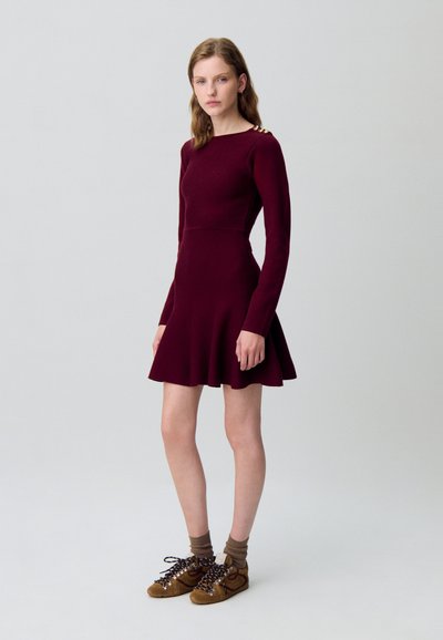 Abito borgogna a maniche lunghe con gonna svasata; realizzato in tessuto morbido. Accentuato da bottoni dorati sulla spalla. Abbinato a sneakers marroni e calze.