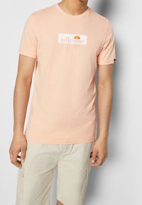 Meruňková bavlněná trička s krátkými rukávy a kulatým výstřihem. Obsahuje bílé obdélníkové logo s multicolor sluneční grafikou.