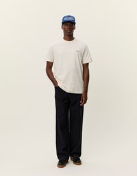 Jasno beżowa koszulka z okrągłym dekoltem, zestawiona z granatowymi spodniami o szerokich nogawkach. Model nosi niebieską czapkę z białym napisem i czarne buty.